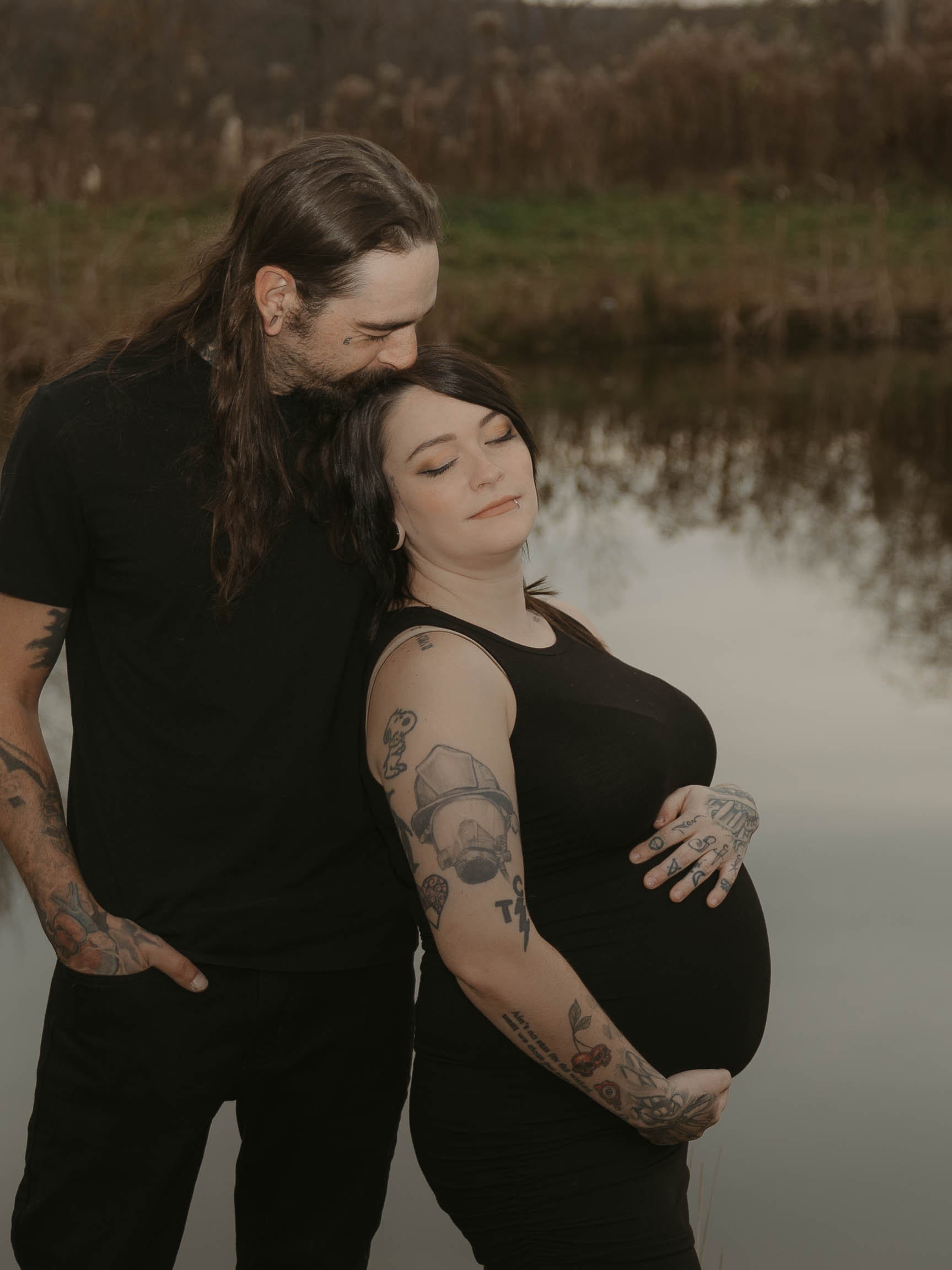 A highlighted image of a couple standing at their pond. She is embracing her growing belly, while he is kissing her head. Both stand in a slow moment embracing this new chapter of life.